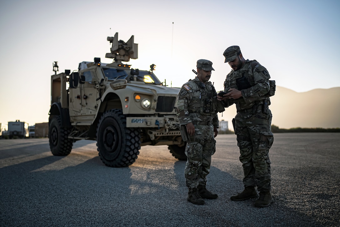 Two soldiers, one holding a small device, talk while standing near a parked military vehicle, with the sun, mountains and other vehicles in the distance.
