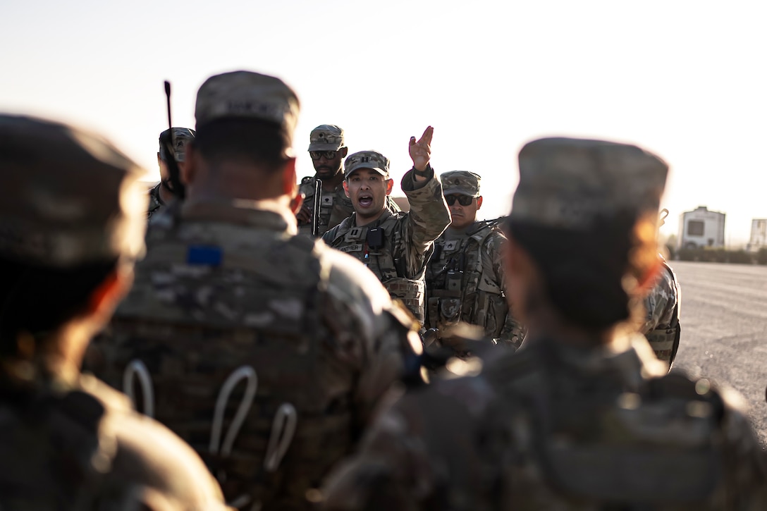 National Guardsmen stand around a soldier giving orders at an outdoor area with two small structures in the background.