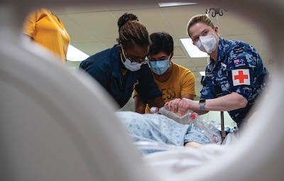 Medical professionals provide intensive care
unit skills training. Three individuals, wearing face masks and protective clothing, including one in an Australian Air Force uniform with a red cross patch, work together using a manual resuscitator. The image is framed from a low angle, possibly through medical equipment, emphasizing the urgency and teamwork in a clinical setting.