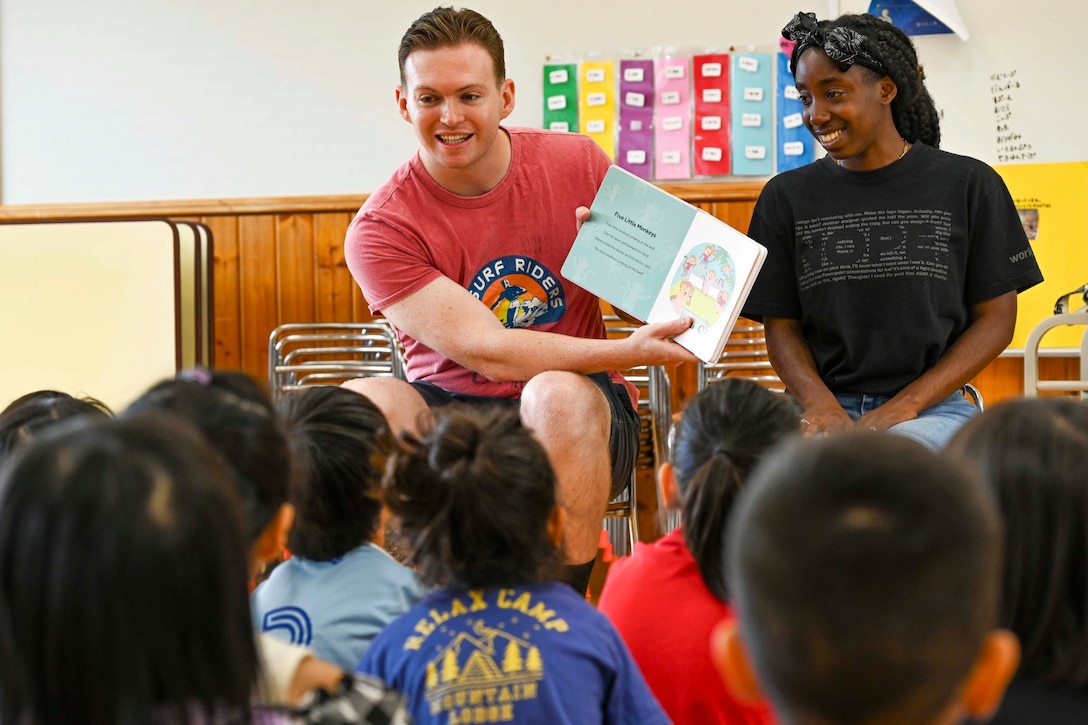 Two seated sailors dressed in casual clothes read a book to kindergarten-age children sitting on the floor in a classroom.