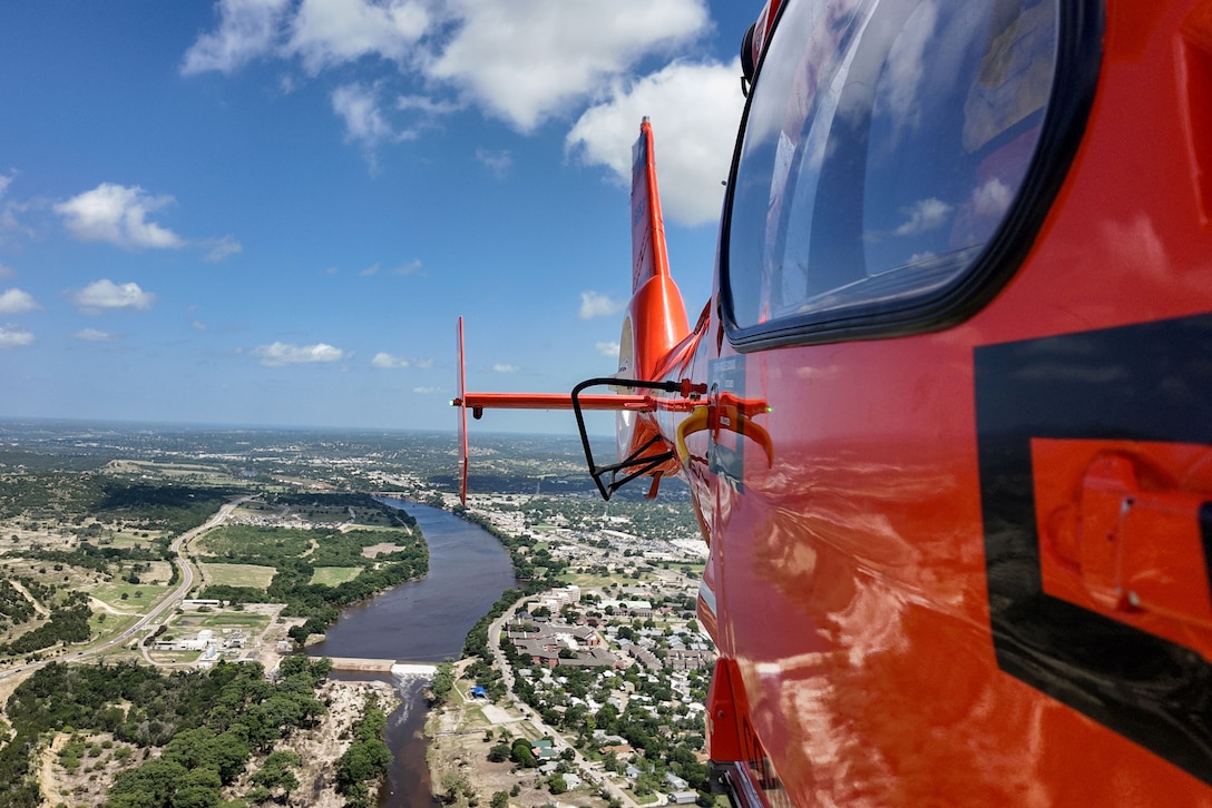 An orange helicopter flies above a river flanked by buildings, roads and trees, with clouds and a blue sky in the background.