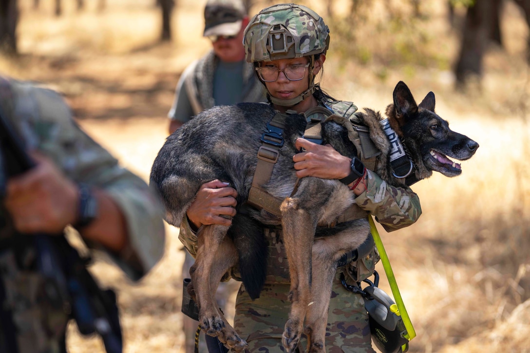 An airman uses both arms to carry a dog in a wooded area, with two other people walking in front and behind the airman.