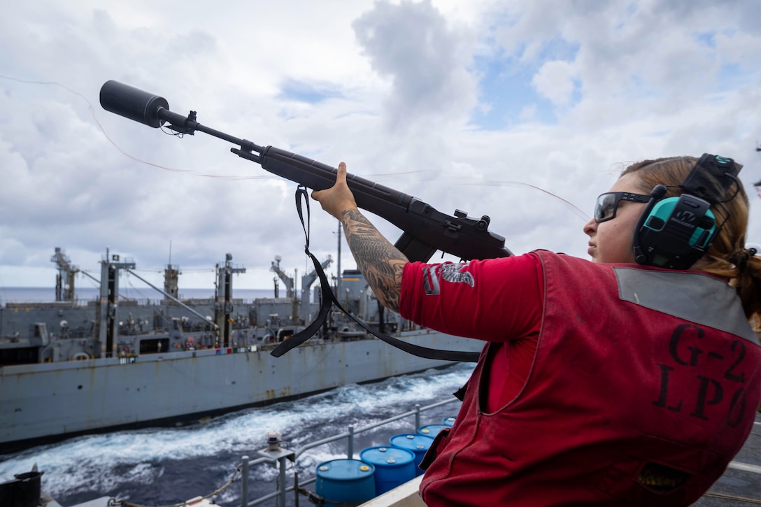 A sailor wearing ear and eye protection fires a shot line from the deck of one ship toward another nearby ship in the ocean on a cloudy day.