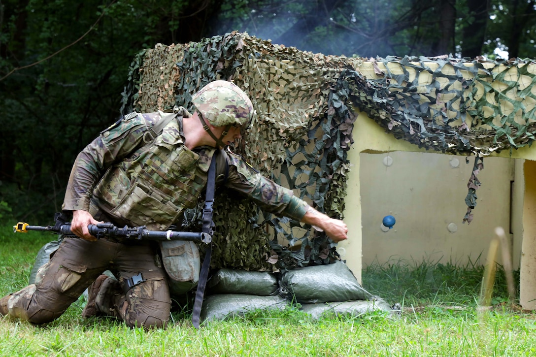 A kneeling soldier tosses a blue hand grenade into a makeshift bunker covered in green netting, with green sandbags stacked near the soldier.