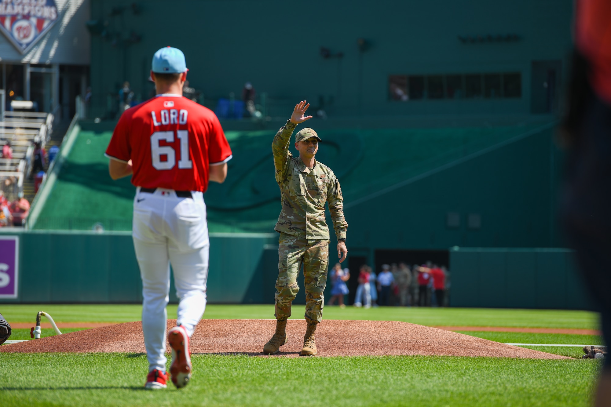 A service member waves from the pitcher's mound of a baseball field.