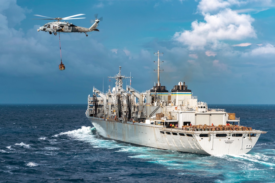 A helicopter with cargo attached to its underside by a heavy line flies away from a ship in the ocean.