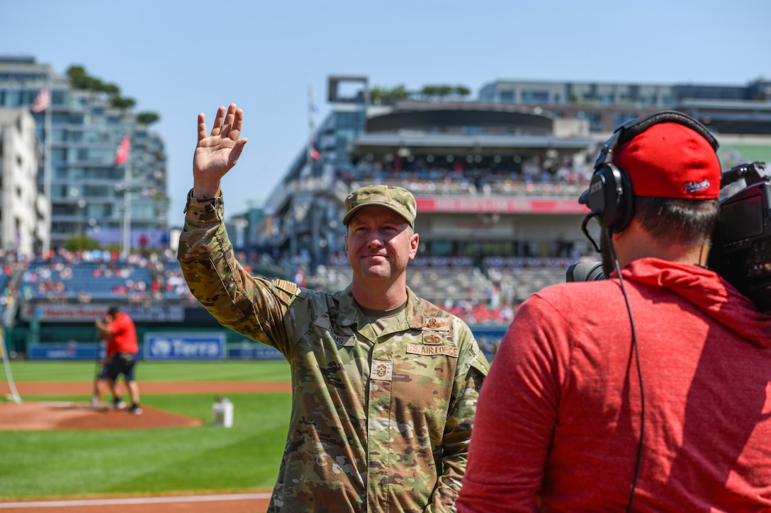 Service member waves at a baseball field.
