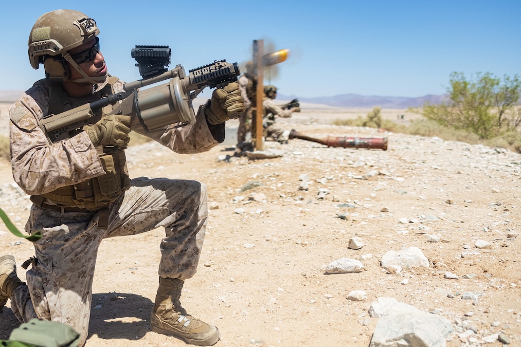 A sailor on one knee fires a grenade launcher in desert-like conditions under a cloudless blue sky.