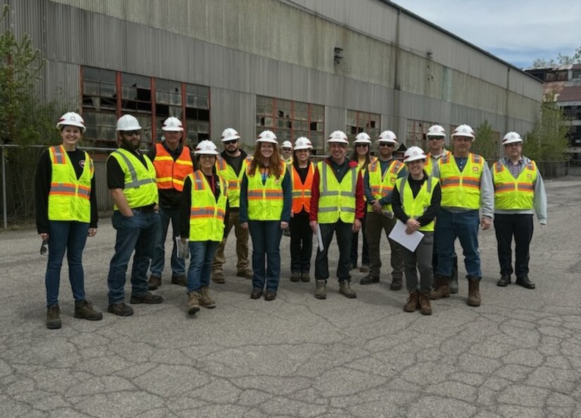 Employees in hard hats and reflective vests pose for a photo in front of a building.