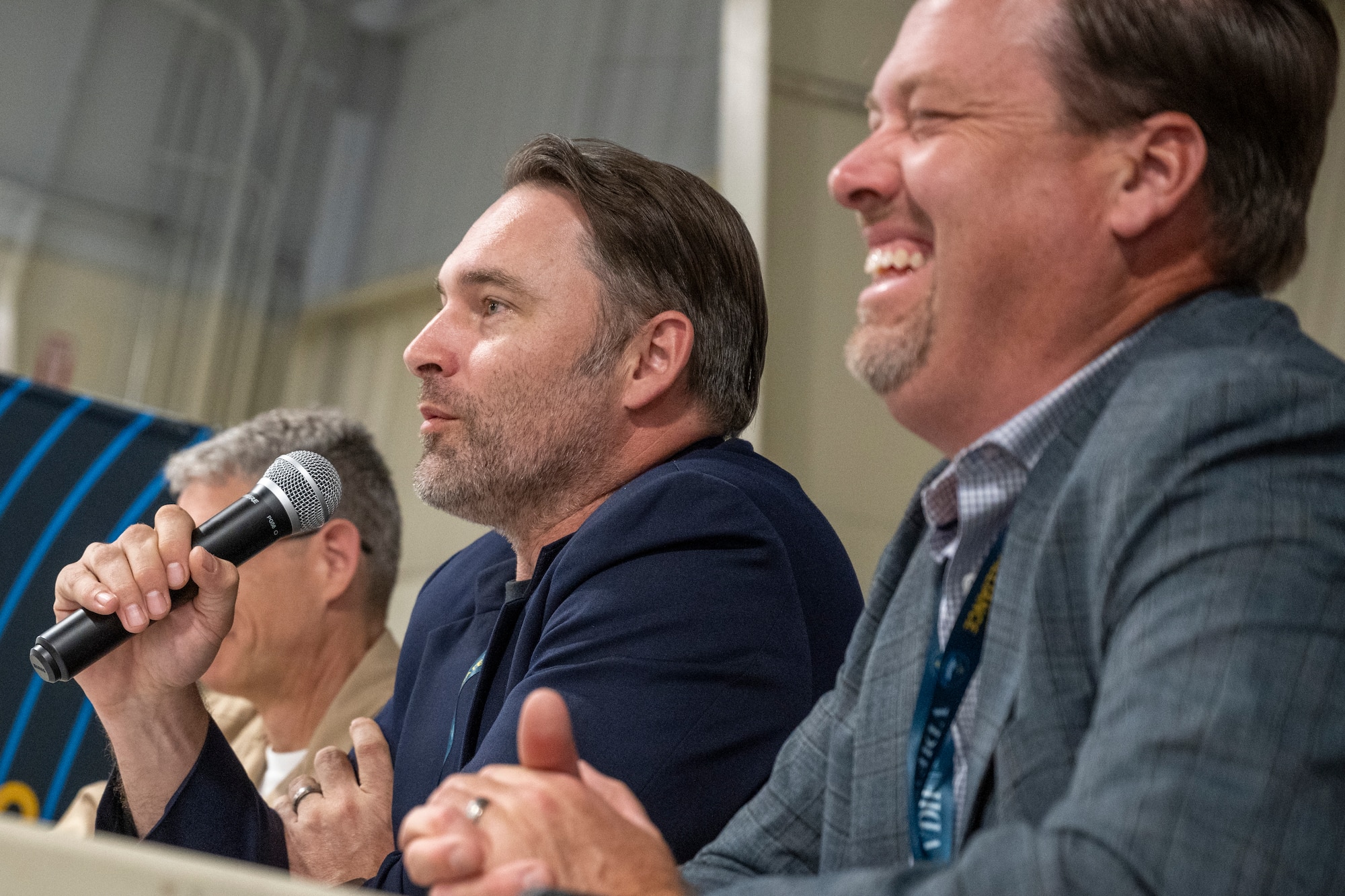 Side view of man speaking on panel, while person to his left laughs
