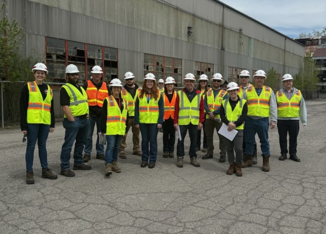 Employees in hard hats and reflective vests pose for a photo in front of a building.