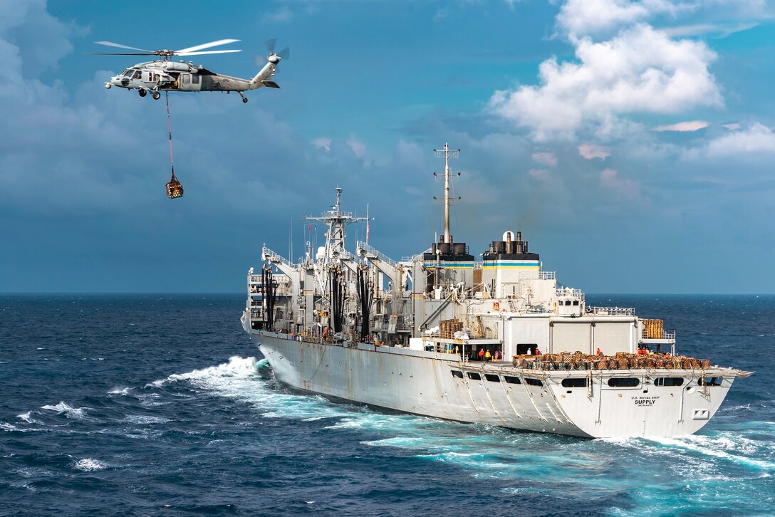 A helicopter with cargo attached to its underside by a heavy line flies away from a ship in the ocean.