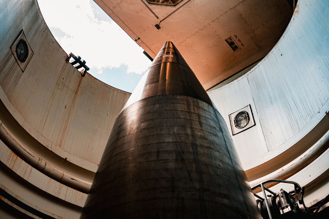 The tip of a missile is seen inside a partially opened silo, with a structure overhead and a sunlit sky in the background.