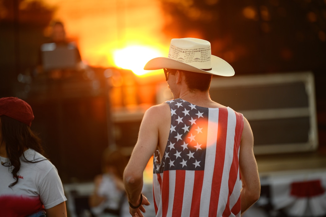 A man in an American flag tank top and cowboy hat stands in front of a sunset.