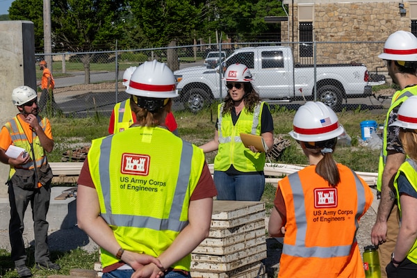 A woman in a yellow safety vest and a hardhat speaks to others in orange and yellow safety vests and hardhats on a dirt ground with a white pickup truck in the background.
