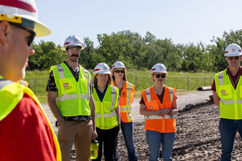 People in orange and yellow safety vests and hardhats stand on brown dirt with green trees and a blue sky in the background.