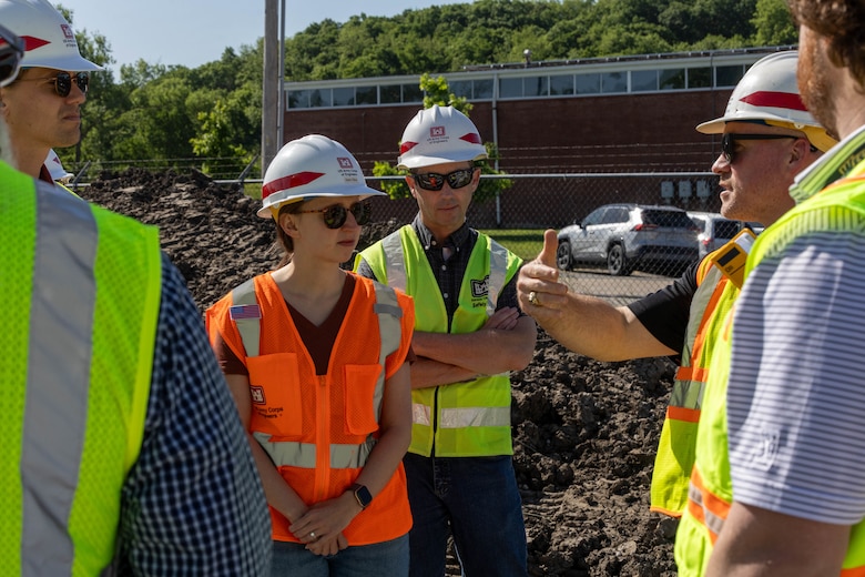 People in orange and yellow safety vests and hardhats stand in a circle and talk with brown dirt and a grey chain link fence in the background.