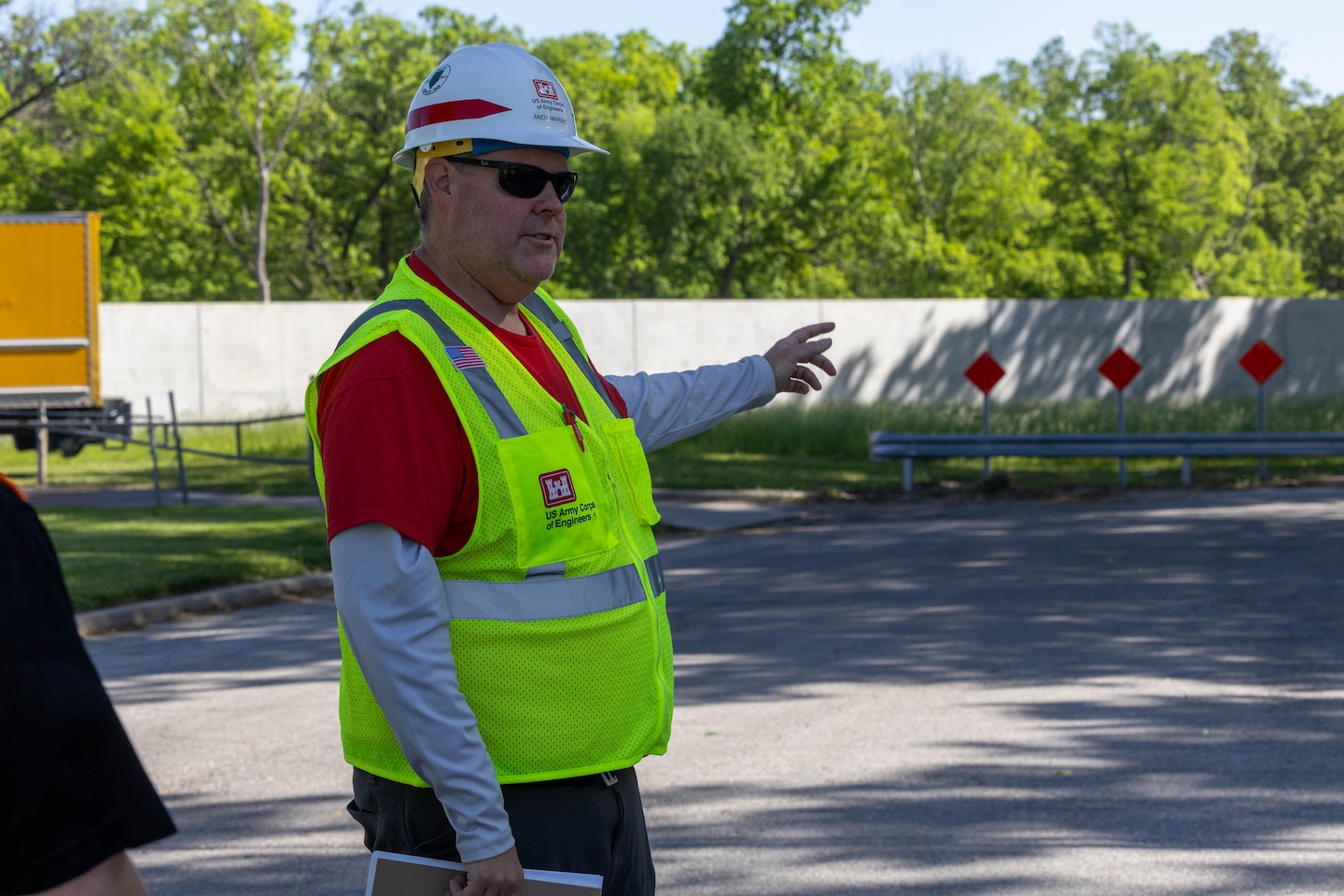 A man in a yellow safety vest and a hardhat points to a grey concrete wall with grey ground in the foreground and green trees in the background.