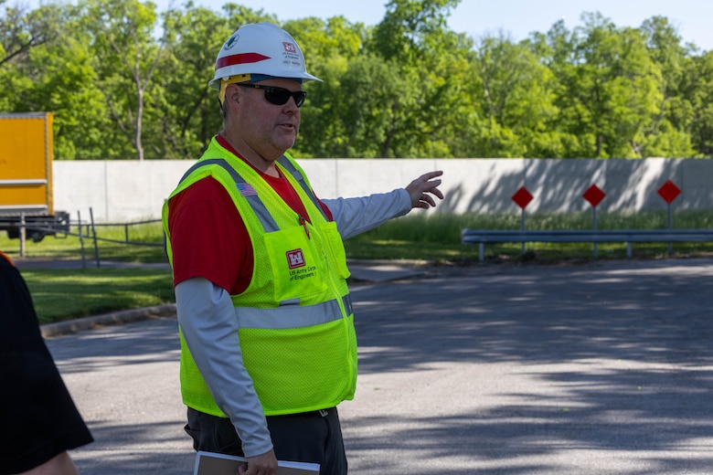 A man in a yellow safety vest and a hardhat points to a grey concrete wall with grey ground in the foreground and green trees in the background.