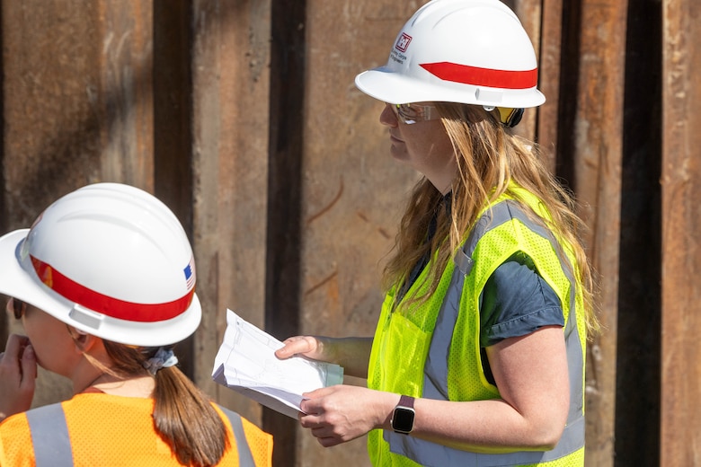 Two women, one in an orange safety vest and one in a yellow safety vest, and both with hardhats stand with brown metal sheets in the background.