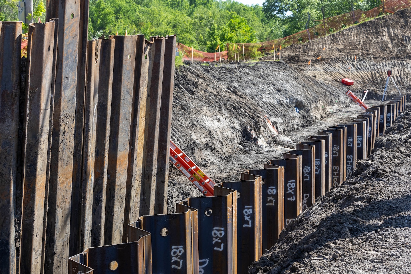 Brown metal components stand in a dirt trench with green trees in the background.