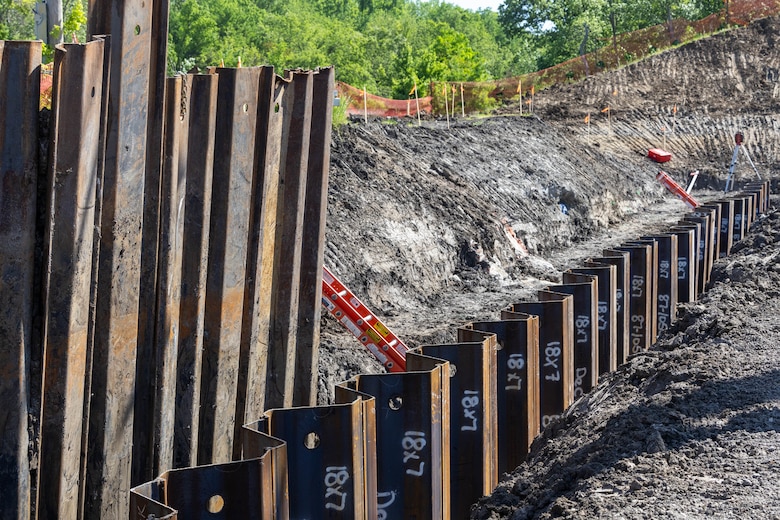 Brown metal components stand in a dirt trench with green trees in the background.