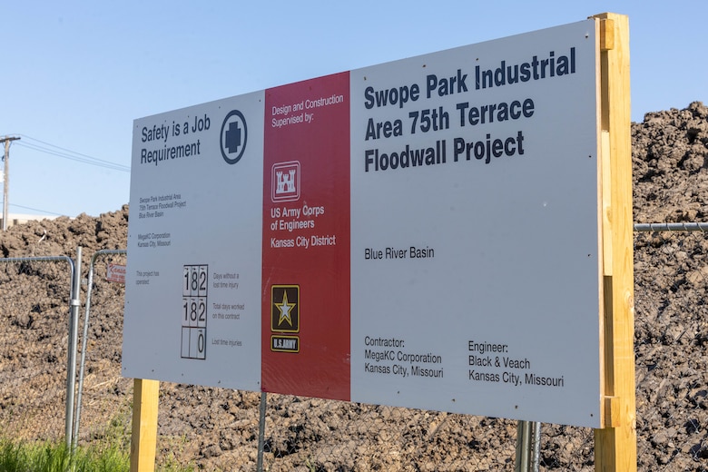 A white and red sign with black text stands in front of a dirt pile and grey chain link fence with blue sky in the background.