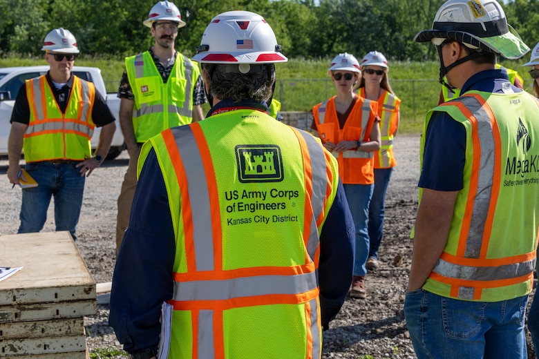People in yellow safety vests and hard hats stand in a circle on dirt with green trees in the background.