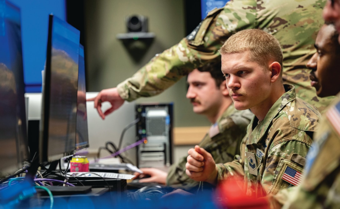 U.S. military personnel in camouflage uniforms work intently at computer stations in a command center. One service member points at a screen while others focus on their monitors. The environment suggests a high-tech operations or cyber defense setting.