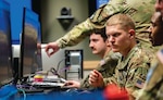 U.S. military personnel in camouflage uniforms work intently at computer stations in a command center. One service member points at a screen while others focus on their monitors. The environment suggests a high-tech operations or cyber defense setting.