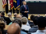 A man in a dark suit stands in a full auditorium as he listens to a question from the audience.