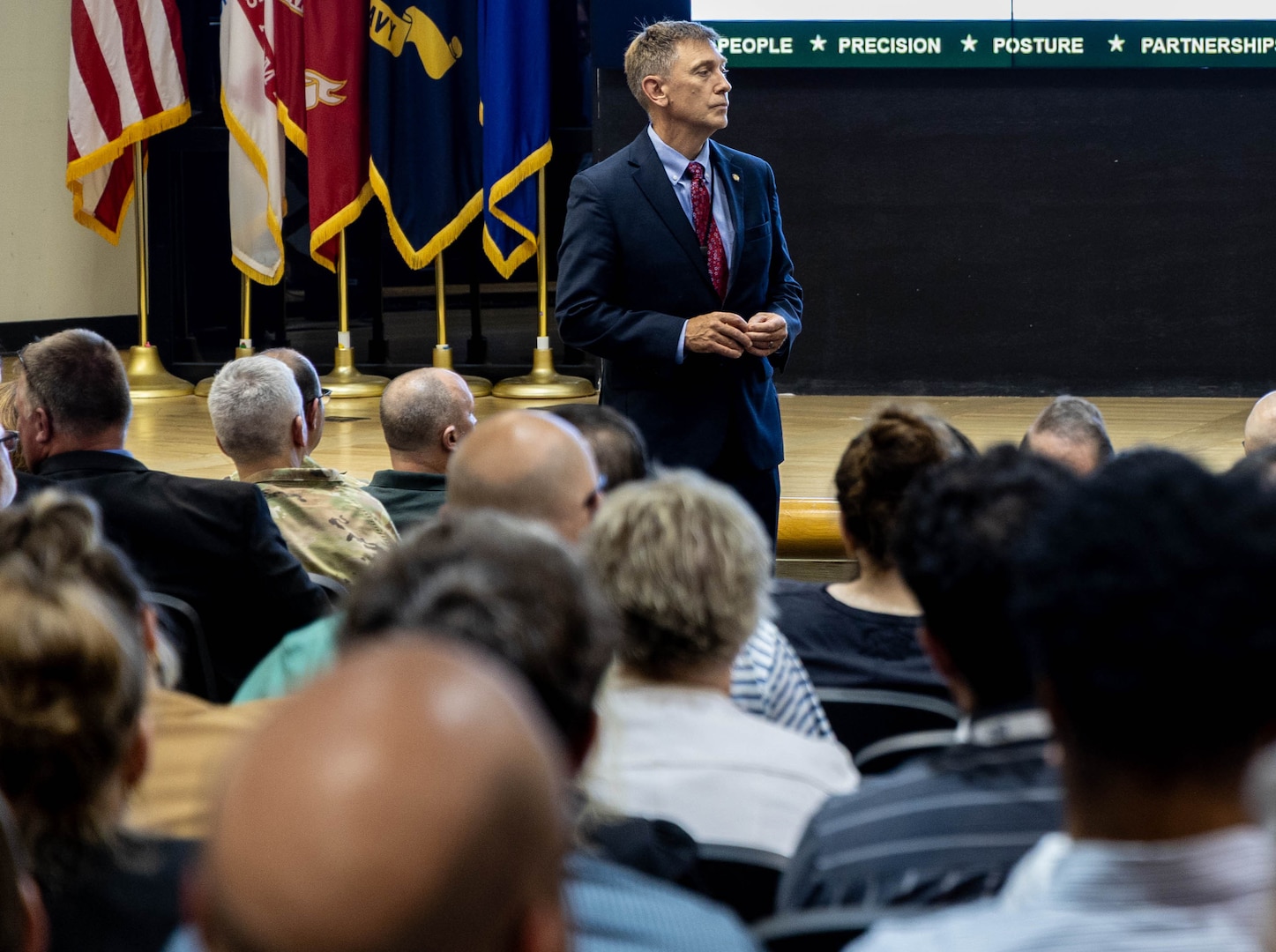 A man in a dark suit stands in a full auditorium as he listens to a question from the audience.