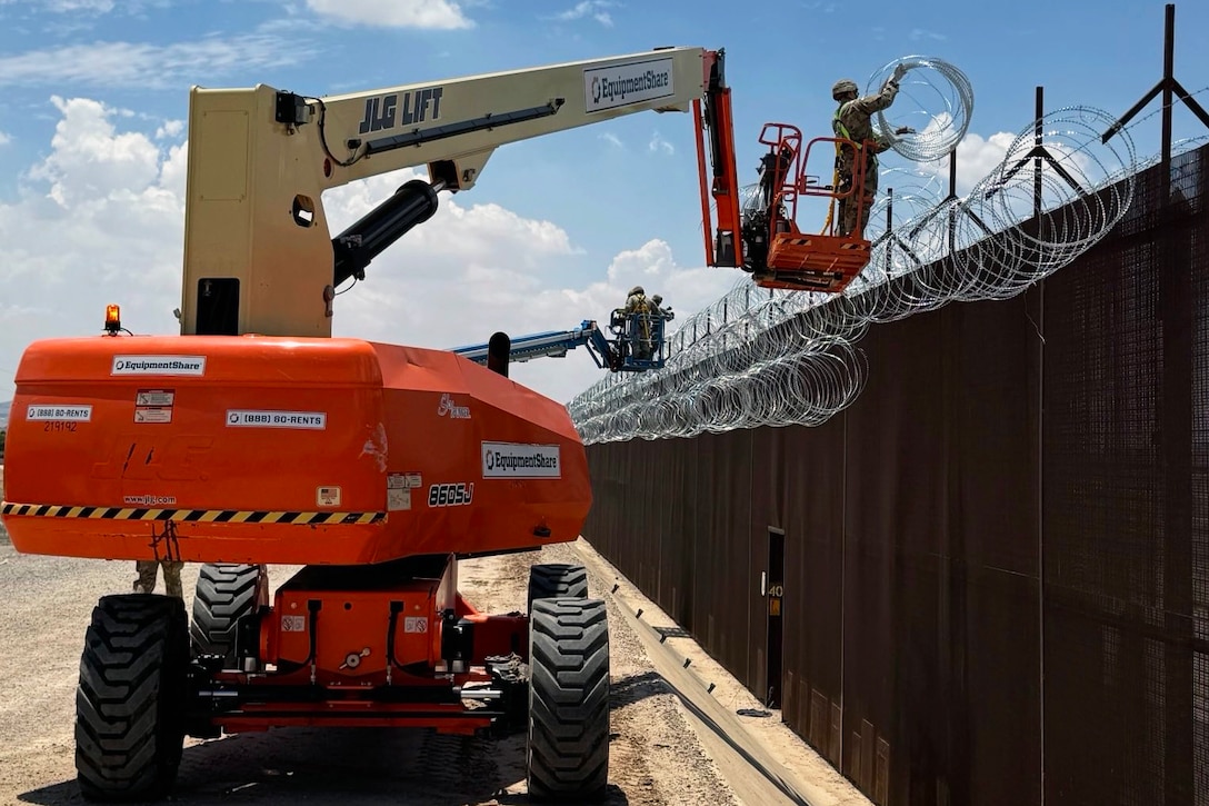 Soldiers standing in elevated work platforms of a boom lift install concertina wire along the top of a tall security barrier.