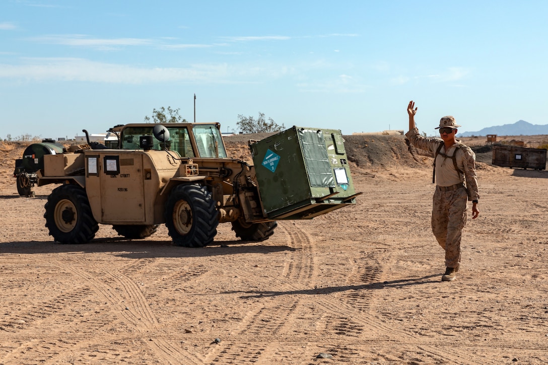 A Marine directs the driver of a rough-terrain forklift transporting cargo at a dirt area outdoors on a sunny day.