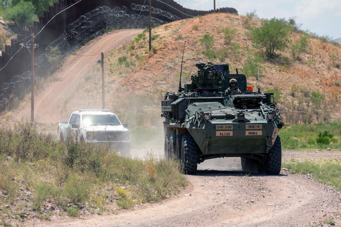 Two soldiers operate an armored vehicle on a dirt road lined on one side by a security barrier in a sparsely vegetated area; a pickup truck follows the armored vehicle.