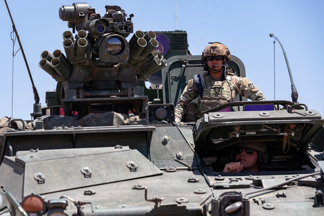 Two soldiers operate an armored vehicle outdoors on a cloudless day.