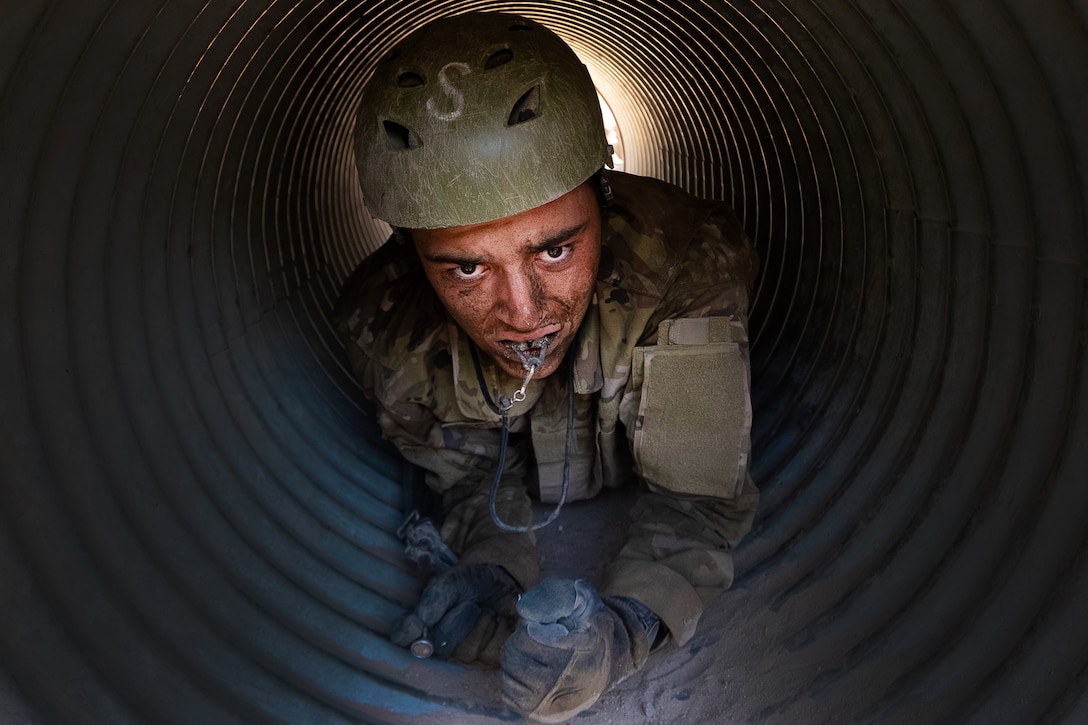 An Air Force cadet trainee wearing a helmet and gloves and clenching an object in the mouth crawls through a tube obstacle.