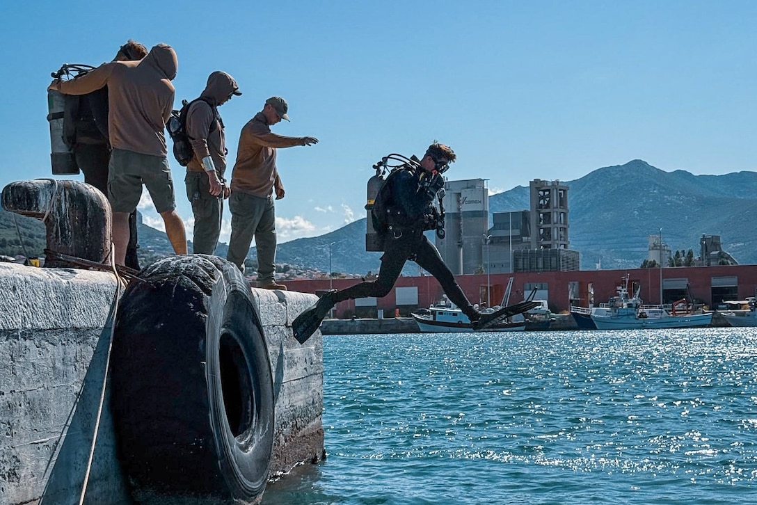 A Navy diver in scuba gear steps from a platform and into a body of water as four others, including one other person in scuba gear, stand on the platform.