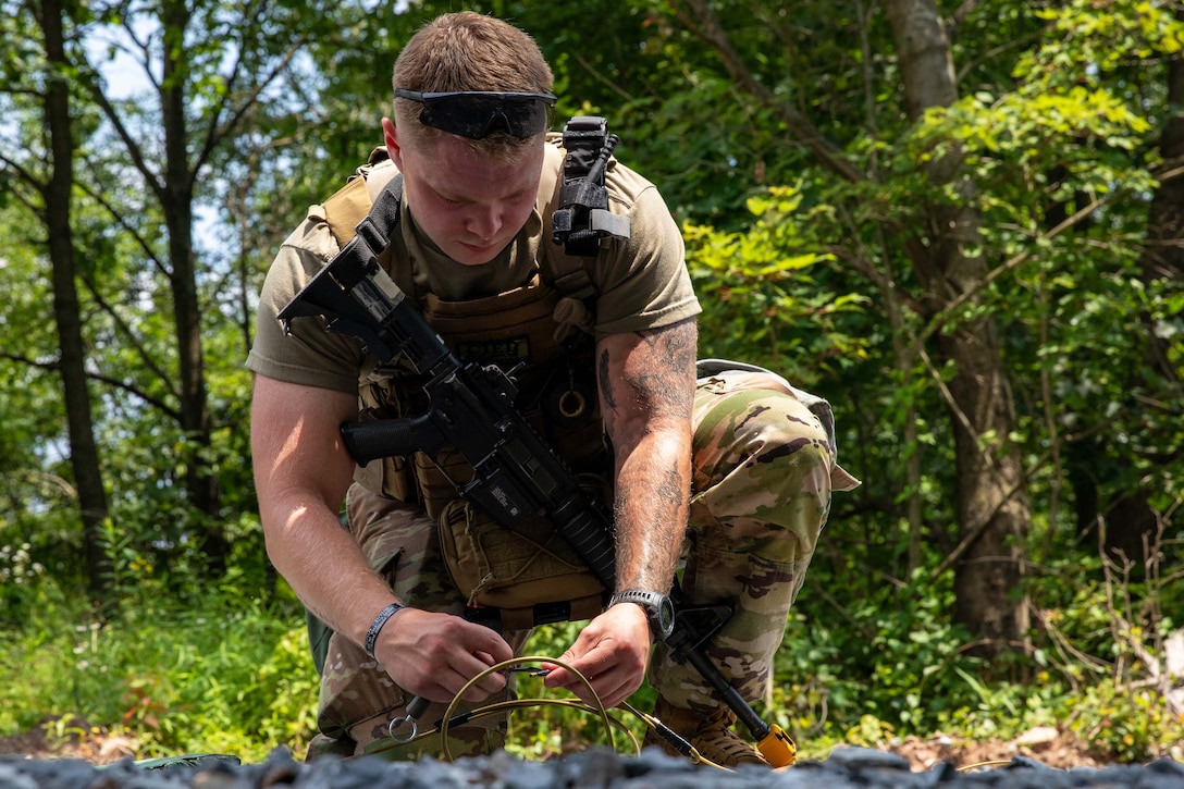 A kneeling soldier attaches cords together on the ground in a wooded area on a sunny day.