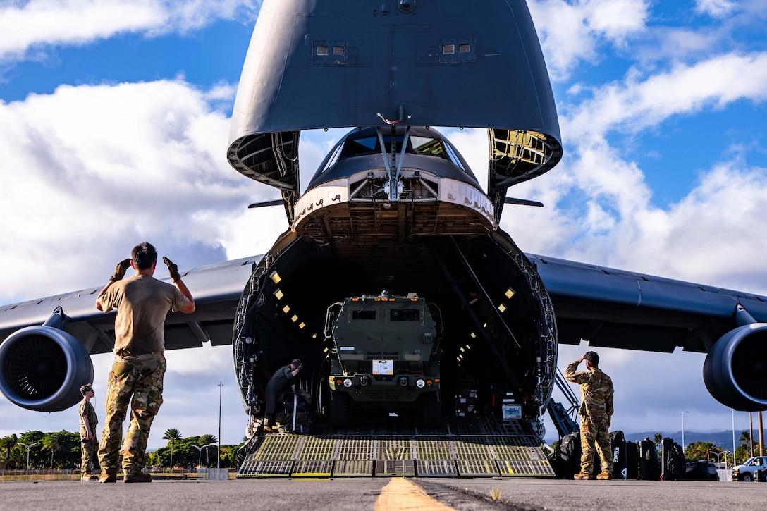 Airmen standing inside and outside the open cargo bay of an airplane unload large equipment on wheels from the aircraft on a runway.