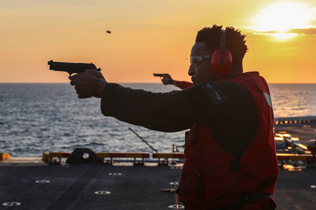 A sailor wearing ear protection fires a pistol from the deck of a ship, with the ocean, the sun and an orange sky in the background.