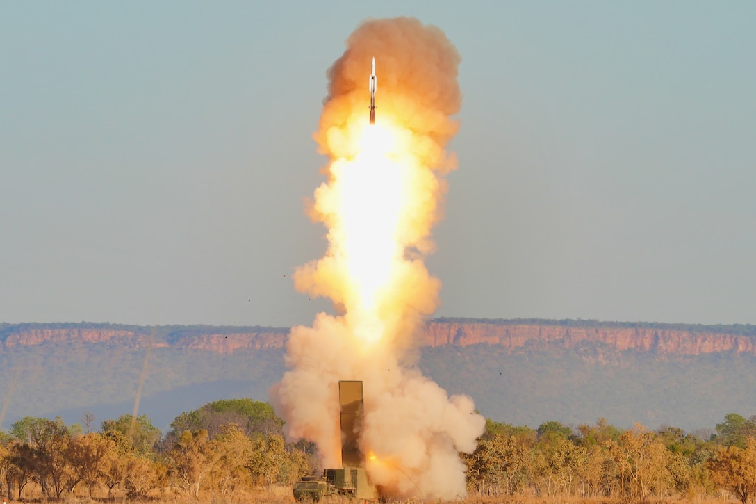 A missile leaves a trail of flames and smoke as it rises from its launch equipment with trees and cliffs in the background.