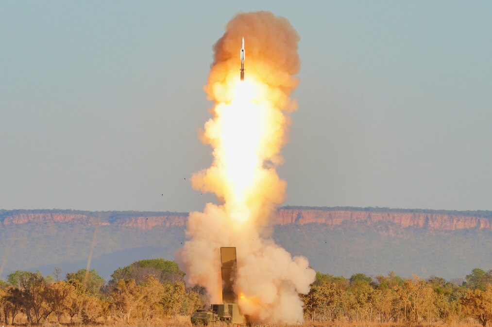 A missile leaves a trail of flames and smoke as it rises from its launch equipment with trees and cliffs in the background.