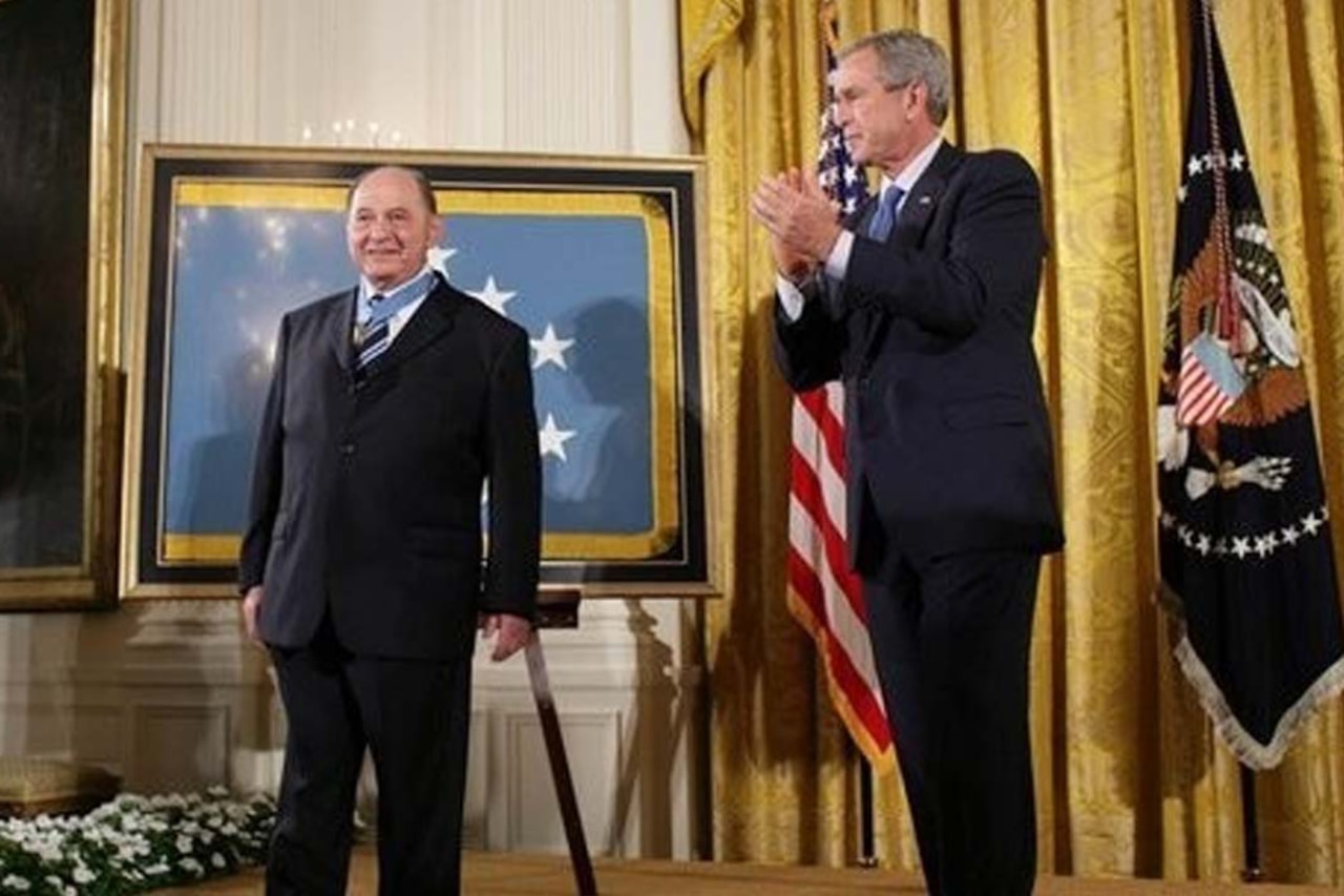 A man in a business suit with a medal around his neck looks outward while another man in a business suit claps beside him. The U.S. and Army flags are displayed behind them.