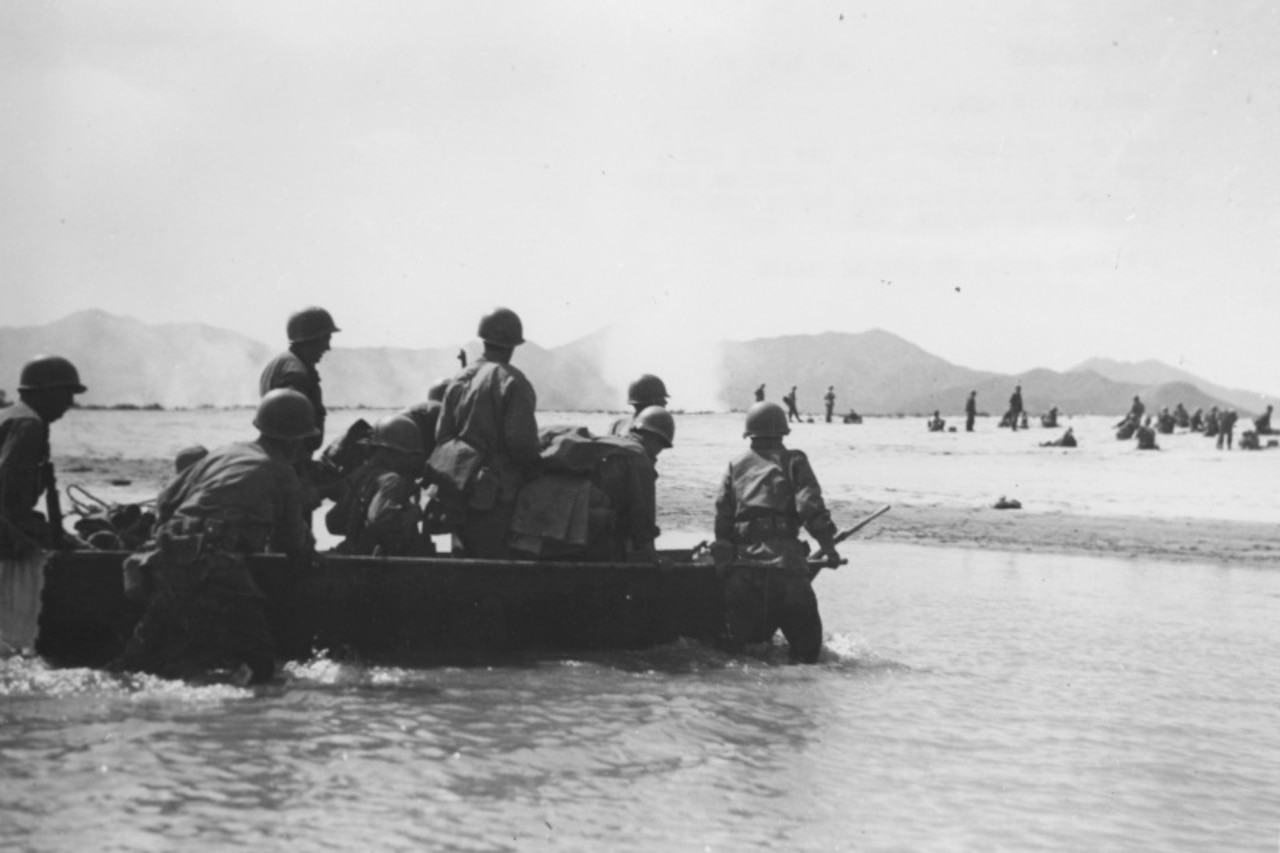 A few men dressed in military uniforms stand in a small boat as others standing in the water push it ashore. Other men are in the background on shore.
