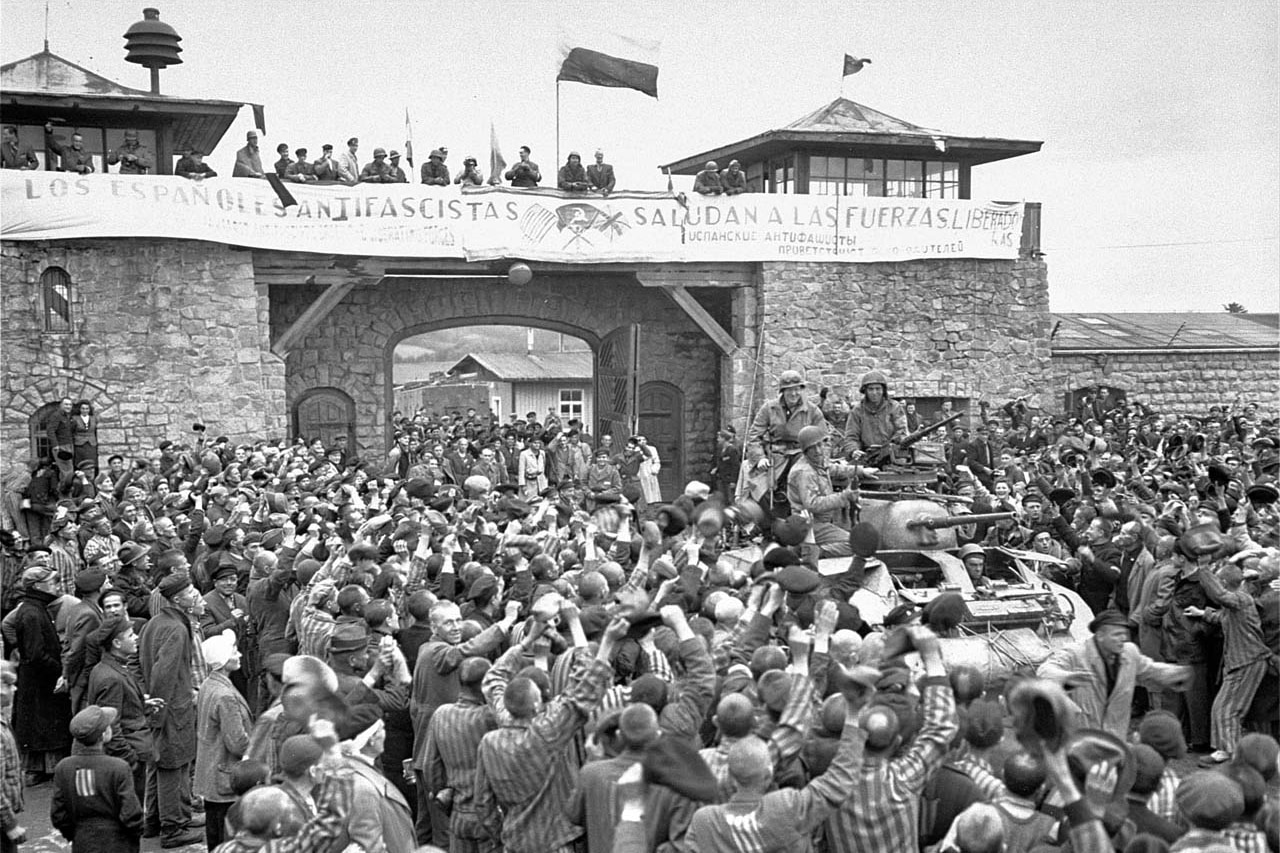 Hundreds of prisoners cheer near stone gates of a concentration camp building as men dressed in military uniforms ride into the camp on a tank.
