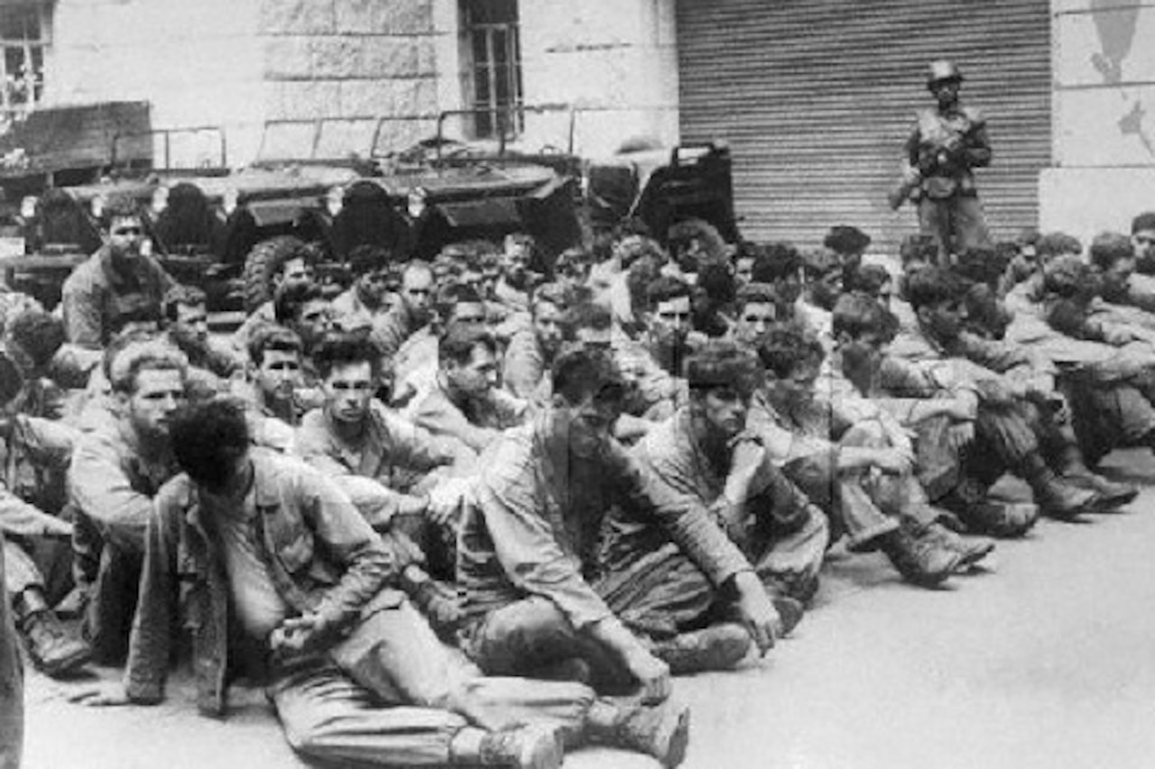 Dozens of emaciated men sit on the ground as a man with a gun wearing a military uniform watches over them.