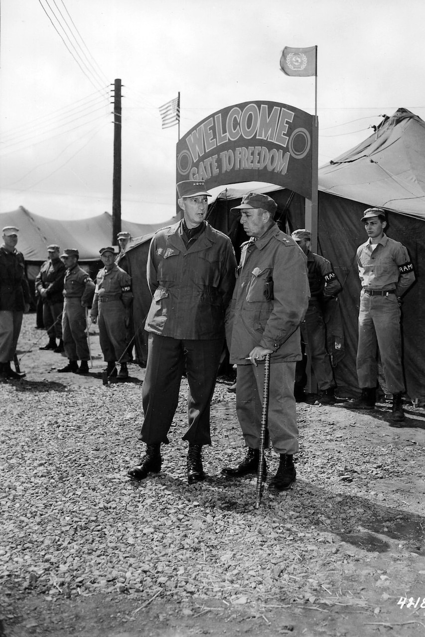 Two men dressed in military uniforms stand in front of a entrance sign that reads "Welcome. Gate to Freedom." Others stand around in the background.
