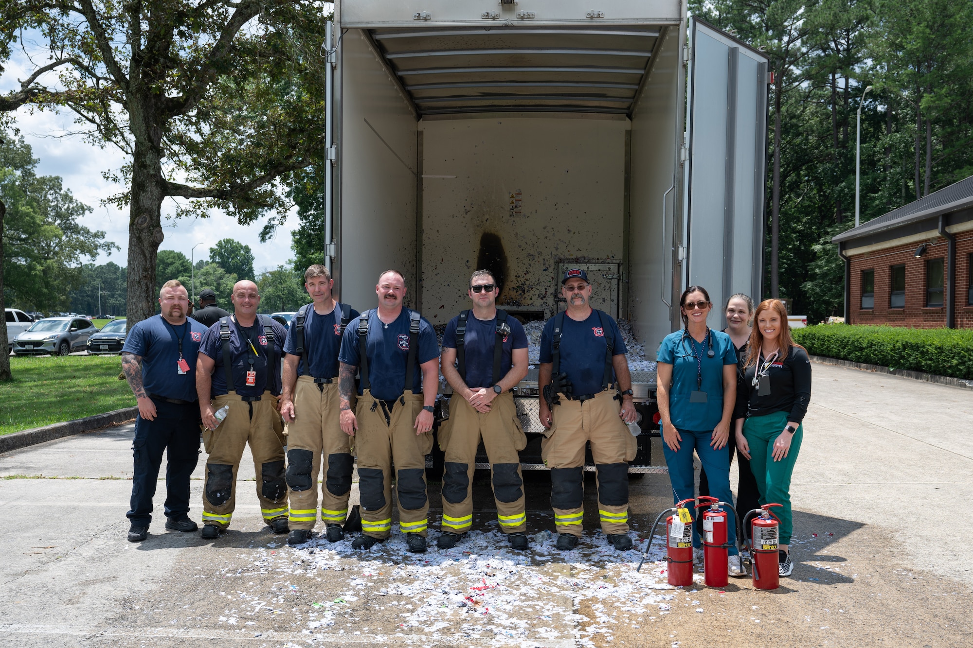 Amanda Van Pelt third from right, Suzanne Hale, second from right, and Kelsey Rob, right, pose for a photo with firefighters from Arnold Air Force Base Fire and Emergency Services after extinguishing a fire outside the Medical Aid Station at Arnold AFB, Tenn., July 11, 2025. Van Pelt, Hale and Rob are staff members with CORE Occupational Medicine, a company subcontracted by the Arnold Engineering Development Complex Test Operations and Sustainment Contractor to provide basic medical services to TOS employees at Arnold AFB, headquarters of AEDC. The CORE team sprang into action when a truck collecting shredded materials unexpectedly caught fire. They used extinguishers to quickly put out the flames, preventing the fire from spreading and minimizing damage. The Arnold FES crew responded to the scene to further douse the fire. (U.S. Air Force photo by Keith Thornburgh) (This image was altered by obscuring badges for security purposes)