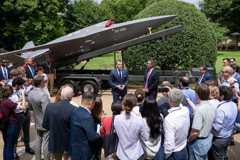 Defense Secretary Pete Hegseth and another civilian stand and talk in front of an unmanned aerial vehicle on display outdoors.